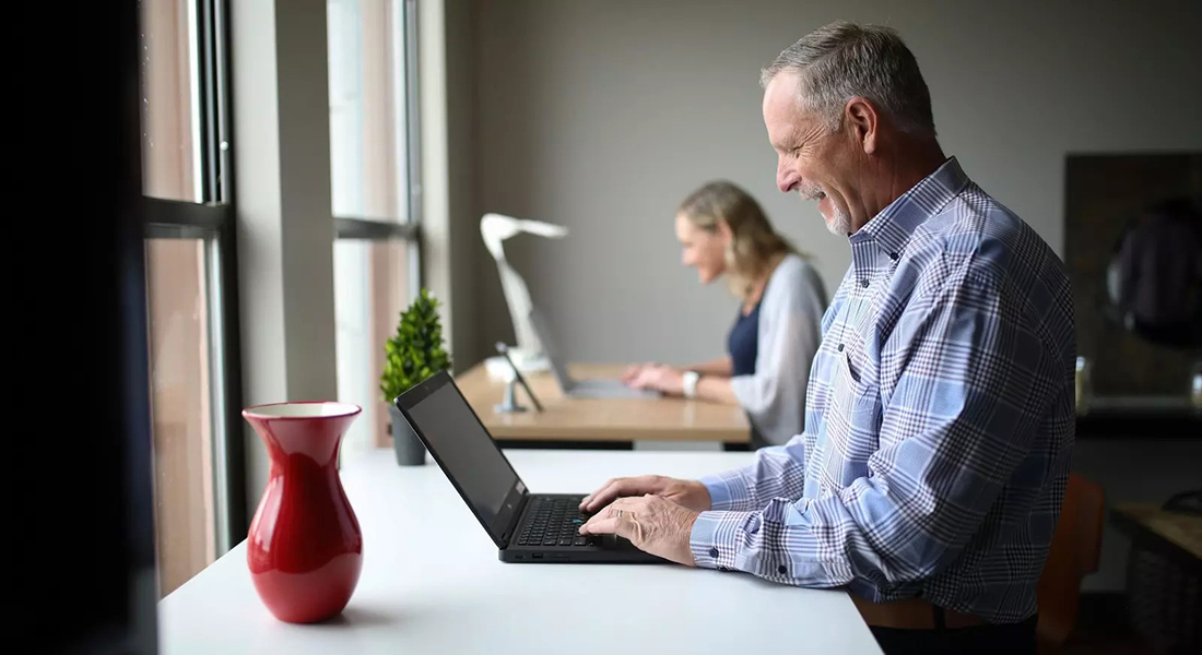 An older and a younger colleague sit next to each other, each with their own laptop.