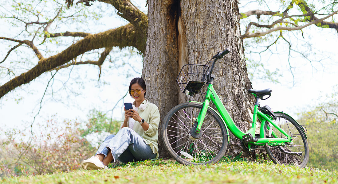 Kvinde der sidder under et stort træ og kigger på en smartphone. Til vores højre for hende står en elcykel.